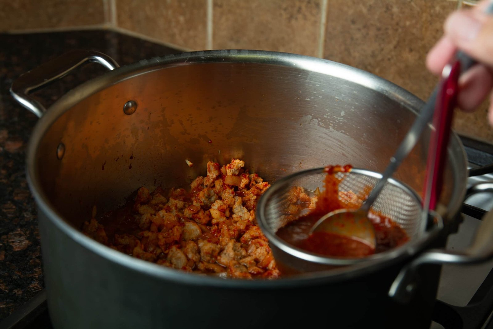 A strainer over the pot of chili with a spoon straining out bits of chili seeds and skin.