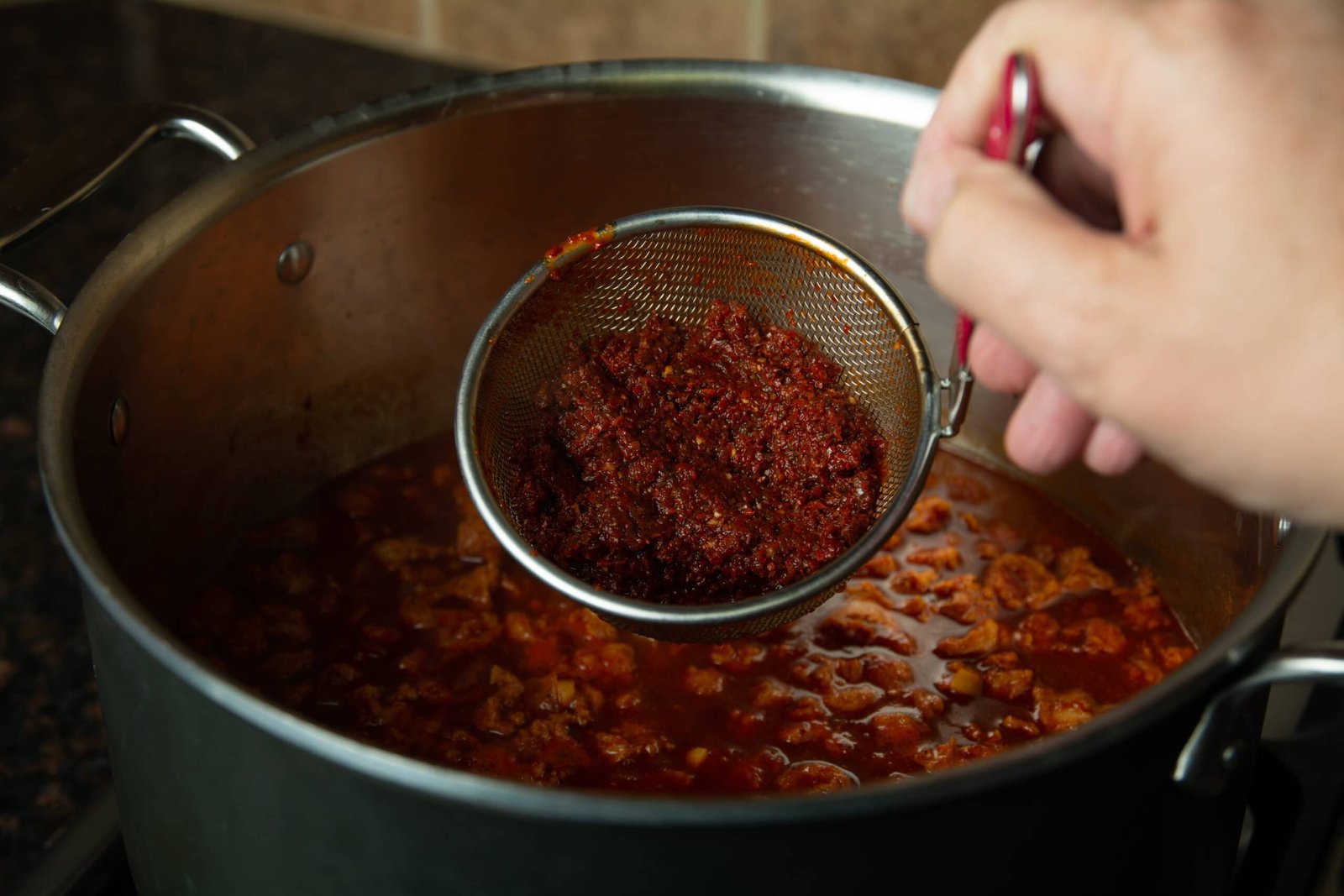 A strainer over the pot of chili with all of the left over bits of chili seeds and skin.