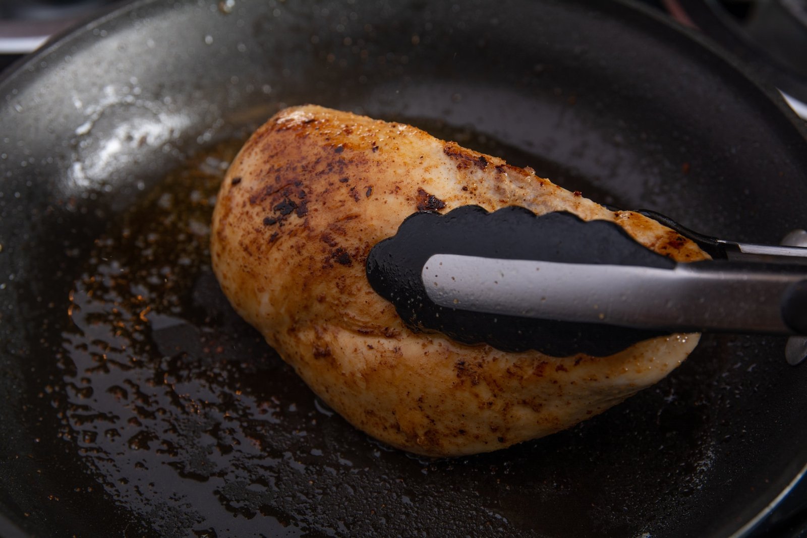 Tongs holding a chicken breast on its side in a pan.