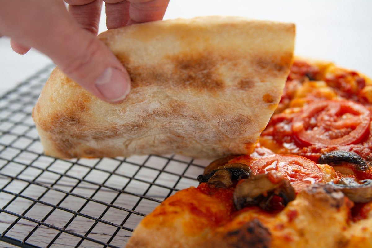 A hand lifting a slice of pizza showing the bottom of the regular crust.