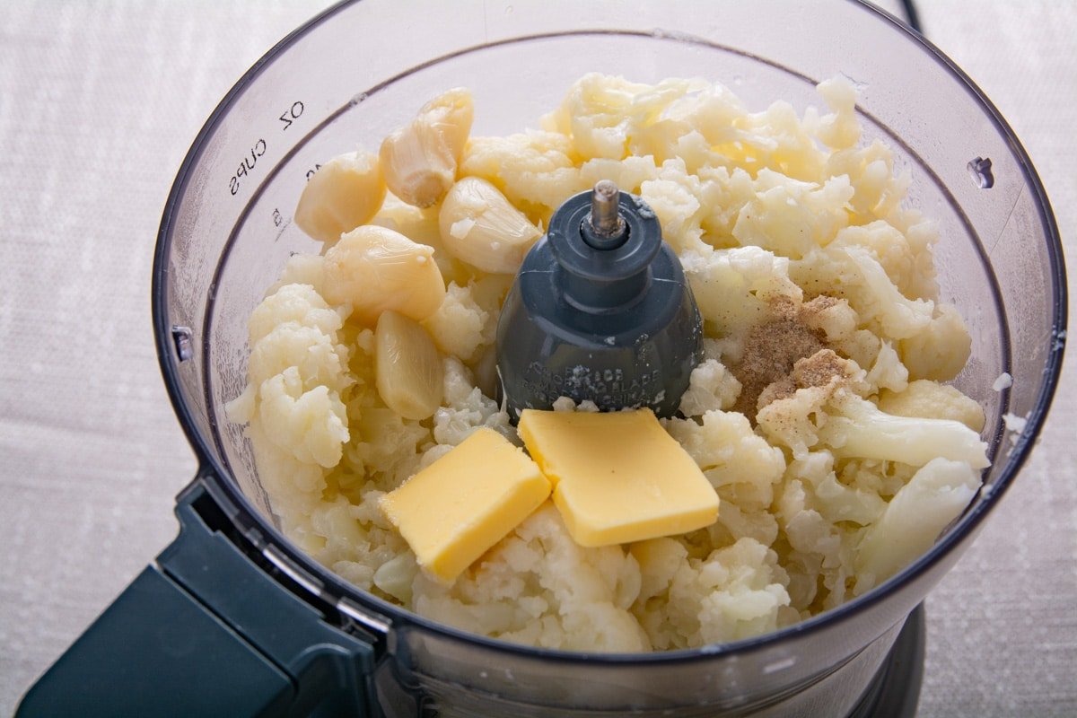 Inside of a food processor with cooked cauliflower, garlic and seasoning.  