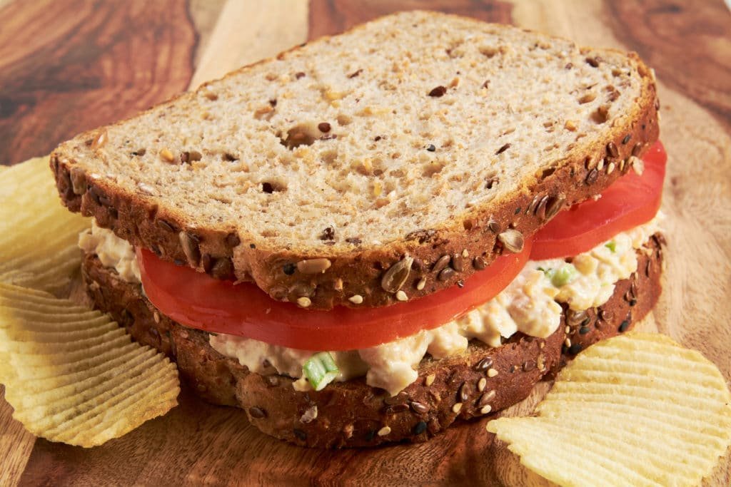 A sandwich made with whole grain bread, tomatoes and chickpea salad sitting on a cutting board.