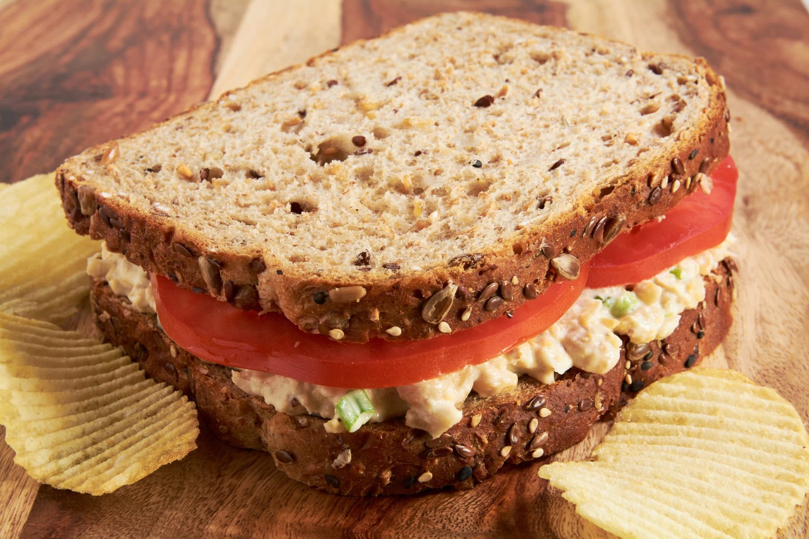 A sandwich made with whole grain bread, tomato slices and chickpea salad on a cutting board.