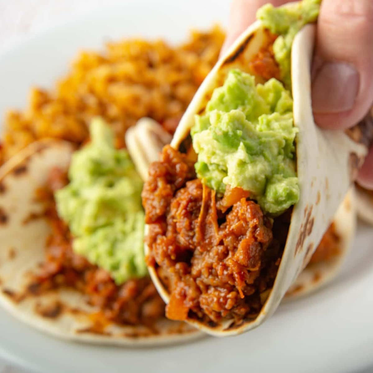 Close up shot of a taco topped with guacamole being held. With more tacos in the background on a plate.