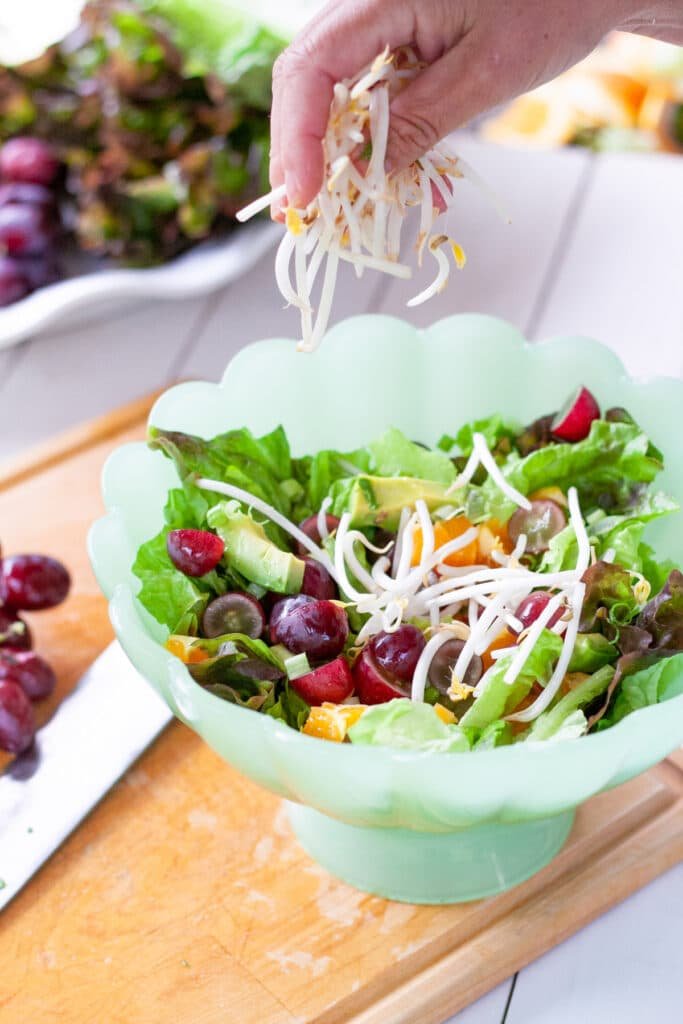 A bowl of salad with a hand sprinkling on bean sprouts.