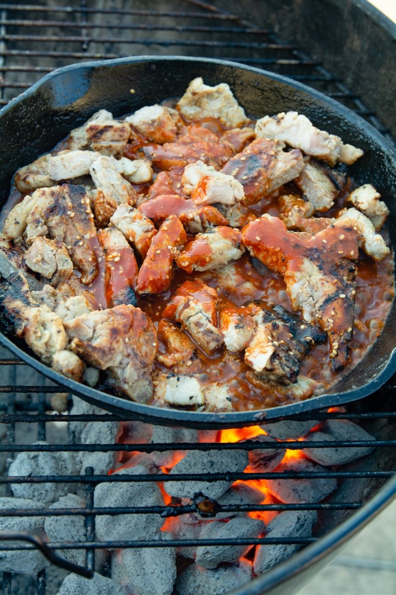 Spicy pork in a cast iron pan cooking on a grill.