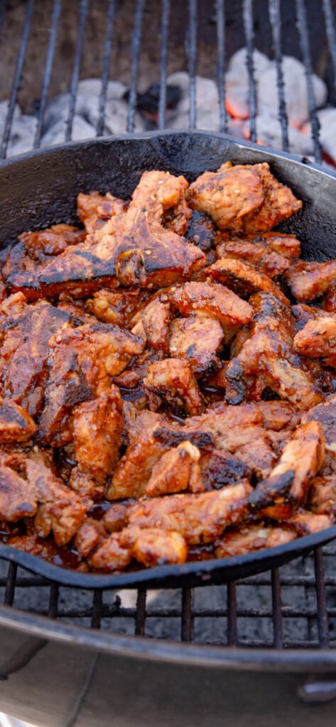 Spicy pork in a cast iron pan cooking on a grill.