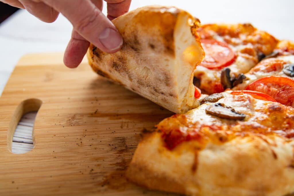 A hand lifting a slice of pizza showing the bottom with black spots of the regular crust.