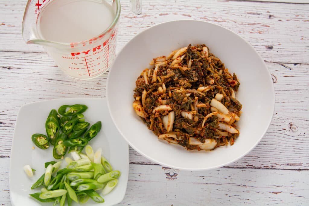 Bowl of seasoned radish leaves, measuring cup of rice water and a plate with green peppers and green onion.