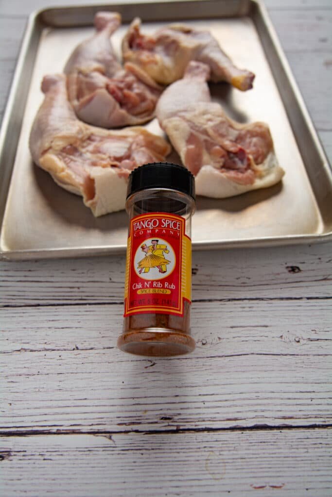 A container of chicken rub and in the background chicken leg quarters