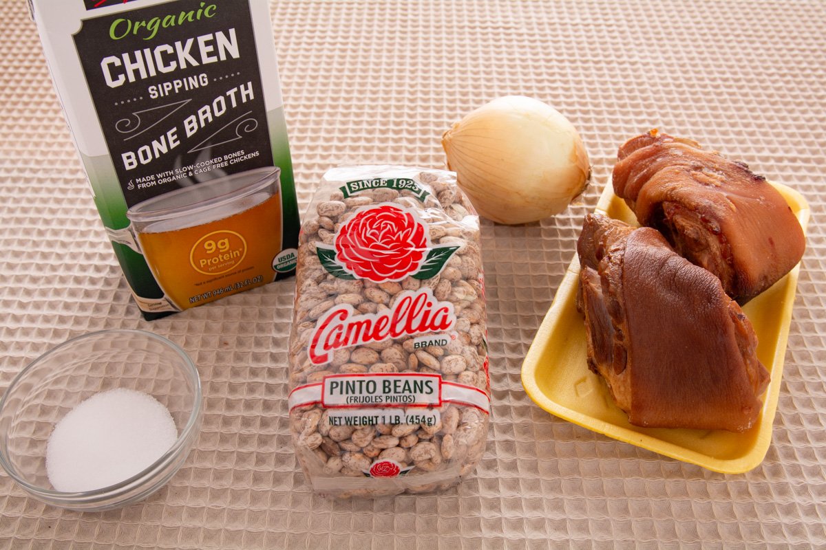 Ingredients on a table, salt, chicken broth, dry pinto beans, onion and ham hocks.